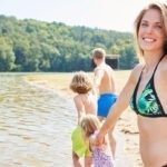 Am Badesee: Junges Paar mit zwei kleinen Kindern am Ufer eines bewaldeten Sees.
